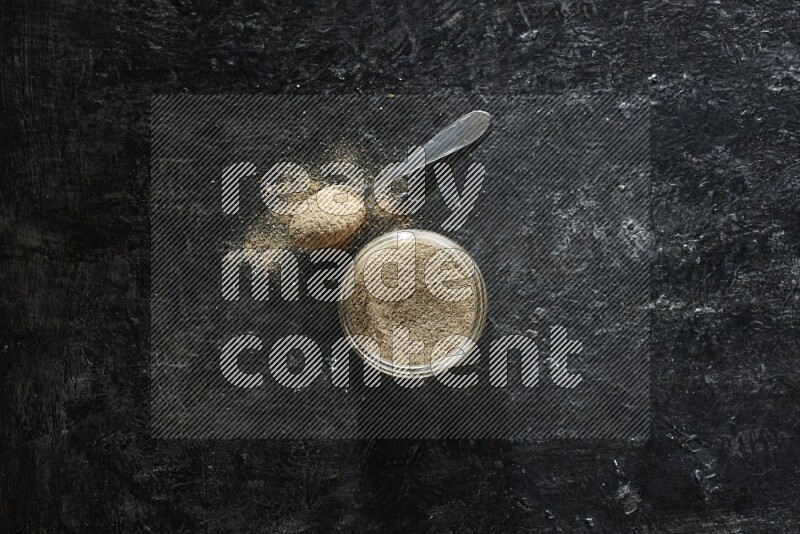 A glass jar and metal spoon full of cardamom powder on textured black flooring