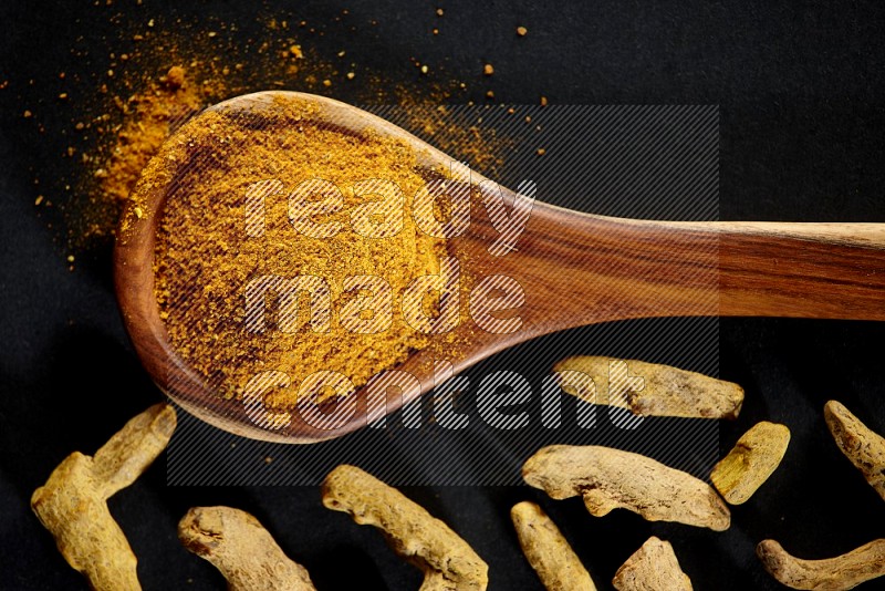 A wooden ladle full of turmeric powder with dried turmeric fingers on black flooring