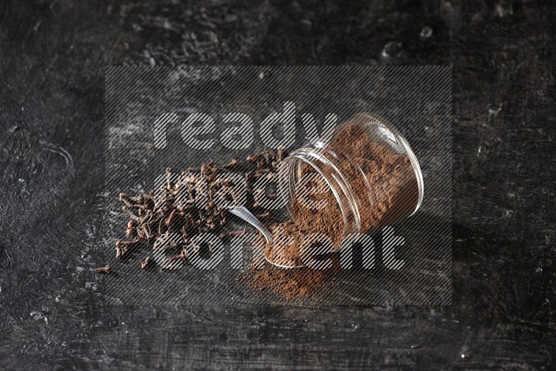 A flipped glass jar and metal spoon full of cloves powder with cloves spread on a textured black flooring