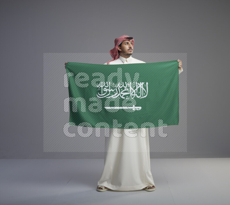 A saudi man standing wearing thob and red shomag holding big saudi flag on gray background