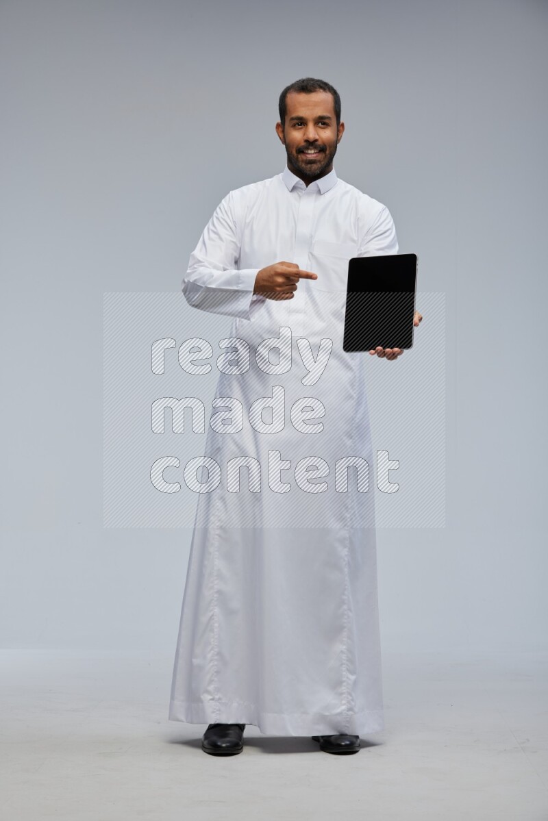 Saudi man wearing Thob standing showing tablet to camera on Gray background