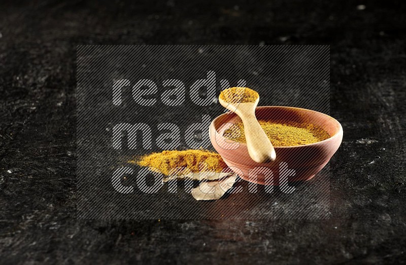 A wooden bowl and a wooden spoon full of turmeric powder on textured black flooring
