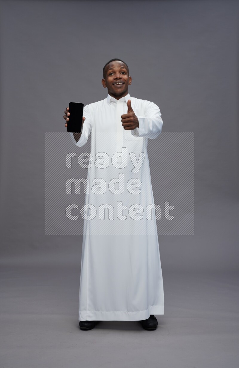 Saudi man wearing thob standing showing phone to camera on gray background
