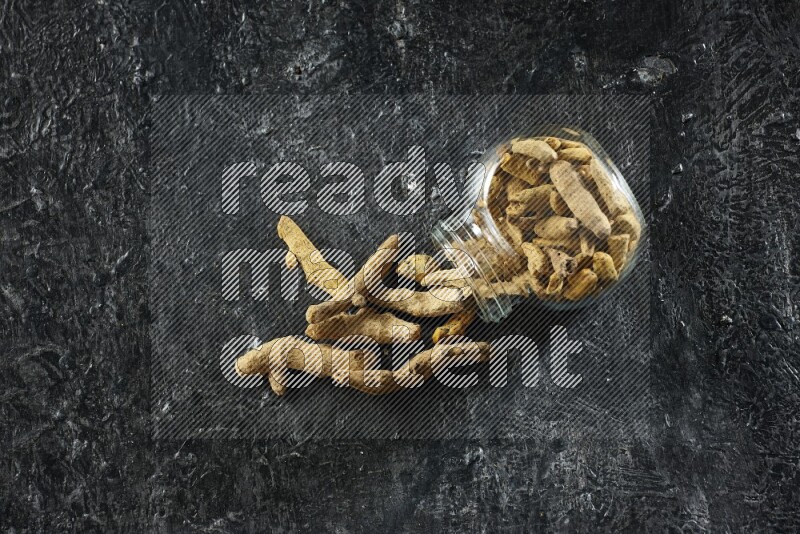 A flipped glass spice jar full of dried turmeric whole fingers and fingers fell out of it on textured black flooring