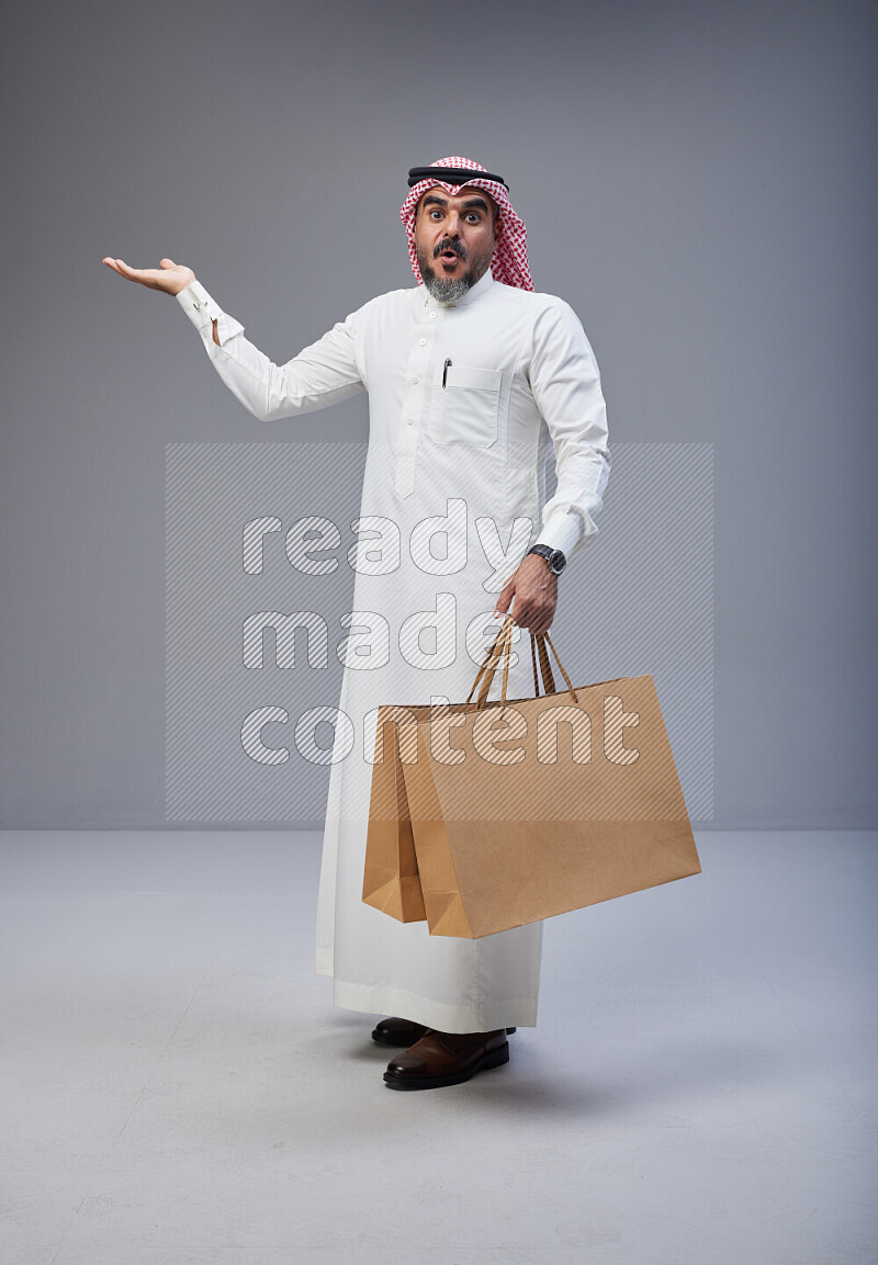 Saudi man Wearing Thob and red Shomag standing holding shopping bag on Gray background