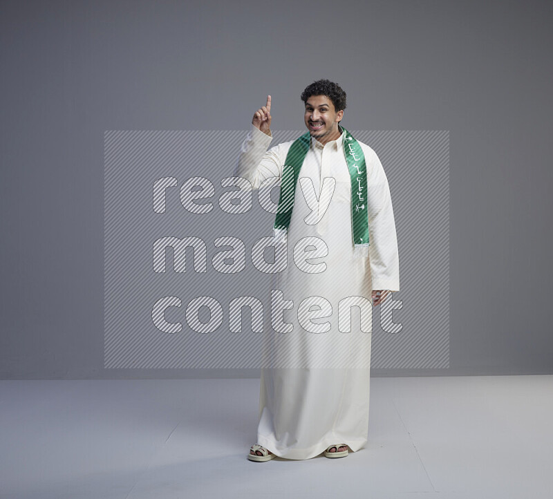 A Saudi man standing wearing thob and Saudi flag scarf on gray background