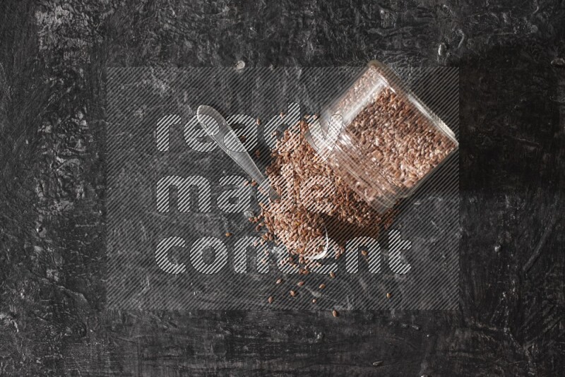 A glass jar full of flaxseeds flipped and seeds spread out with a metal spoon full of the seeds on a textured black flooring