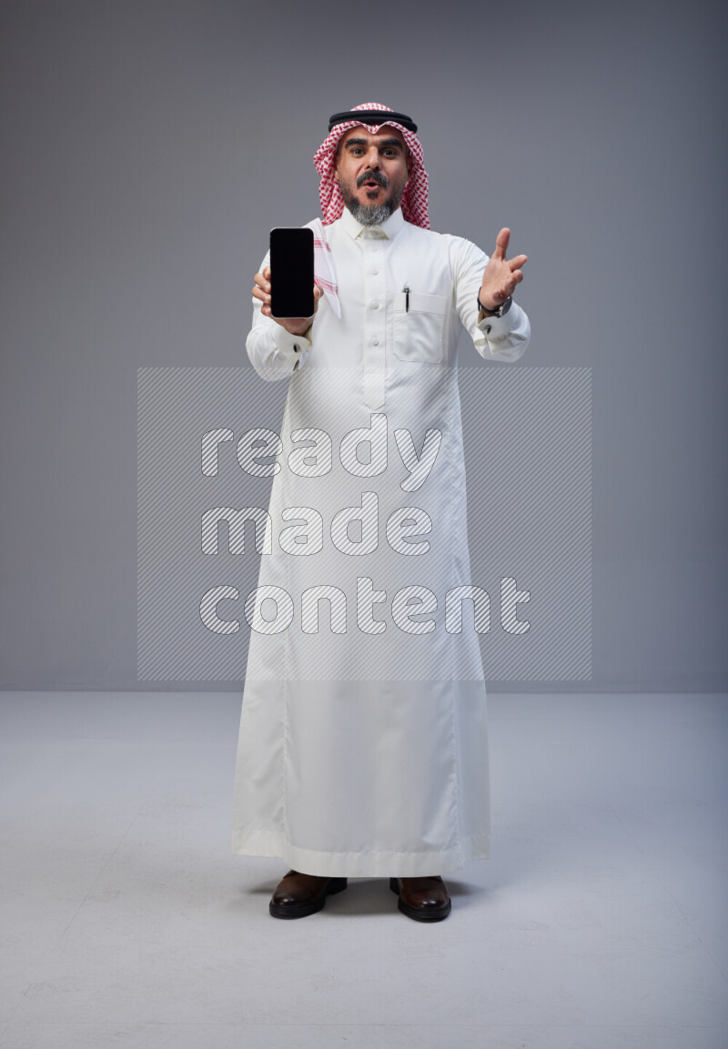 Saudi man Wearing Thob and red Shomag standing showing phone to camera on Gray background