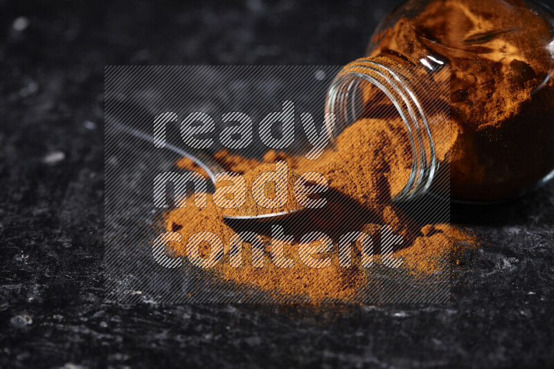 A glass jar full of ground paprika powder flipped with some spilling powder on black background