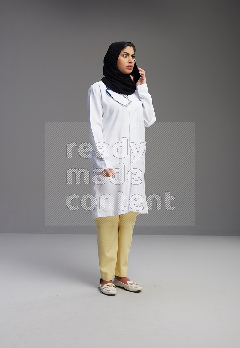 Saudi woman wearing lab coat with stethoscope standing talking on phone on Gray background