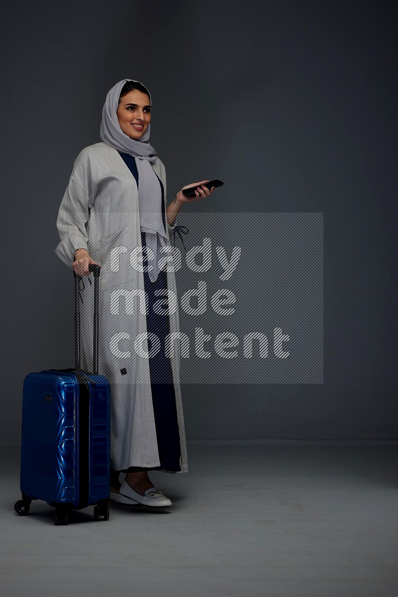 A Saudi woman wearing a light gray Abaya and head scarf standing holding a phone and being shocked  eye level on a grey background