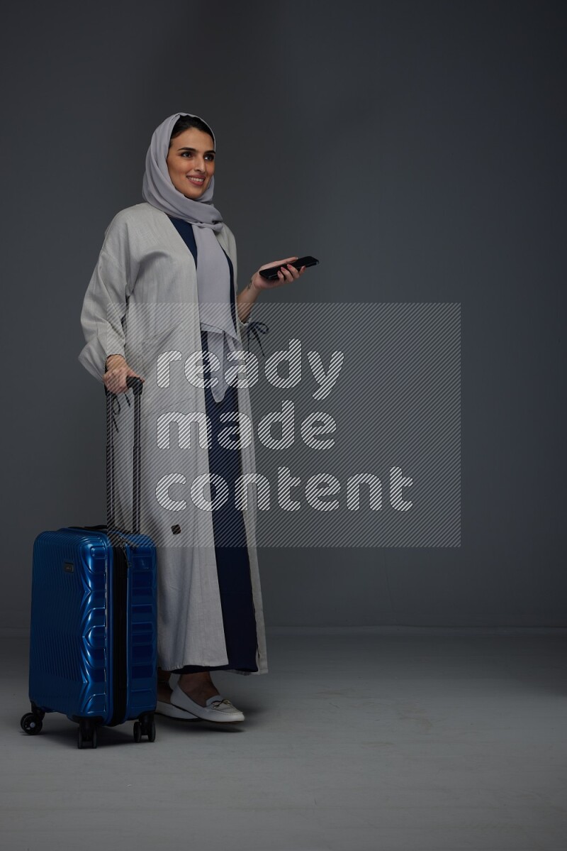 A Saudi woman wearing a light gray Abaya and head scarf standing holding a phone and being shocked  eye level on a grey background