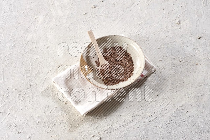 A multicolored pottery plate full of flax seeds with a wooden spoon full of the seeds on a napkin on a textured white flooring