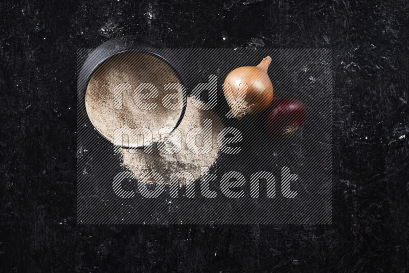 A black pottery bowl full of onion powder with fallen powder from it on black background