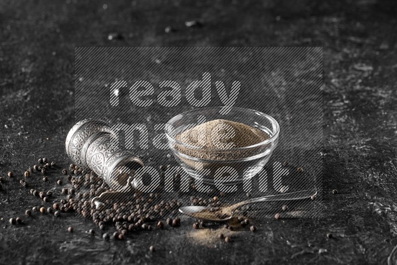 A glass bowl full of black pepper powder with black pepper beads, a turkish metal pepper grinder and a metal spoon on textured black flooring