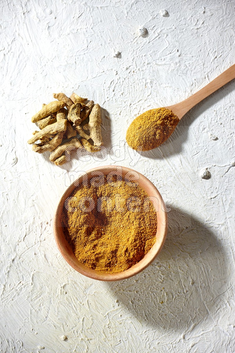 A wooden bowl and wooden spoon full of turmeric powder with dried turmeric fingers on textured white flooring