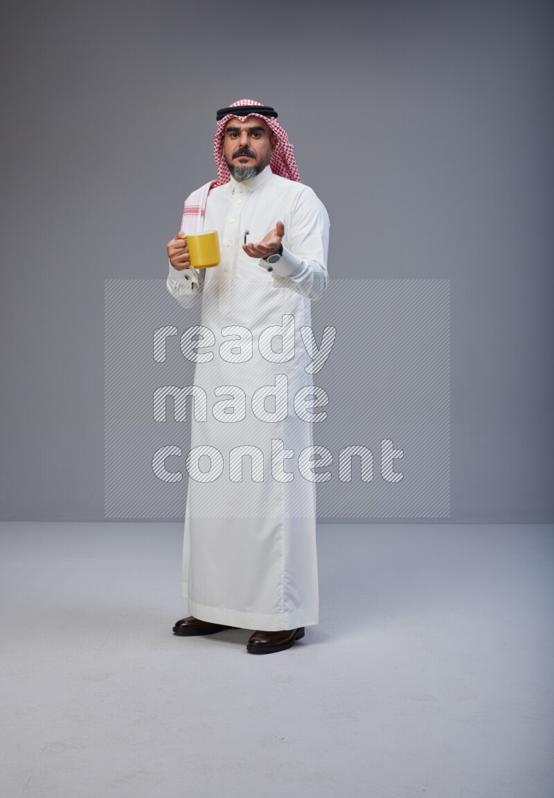 Saudi man Wearing Thob and red Shomag standing holding mug on Gray background