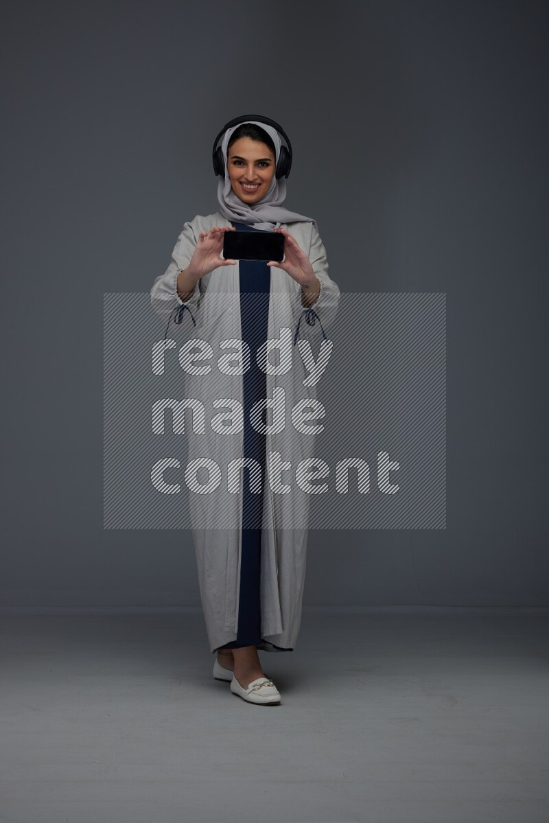 A Saudi woman wearing a light gray Abaya and head scarf setting on a dark grey chair and watching on the tablet eye level on a grey background