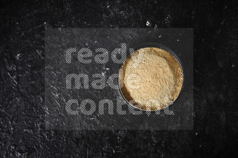 A black pottery bowl full of ground ginger powder on black background