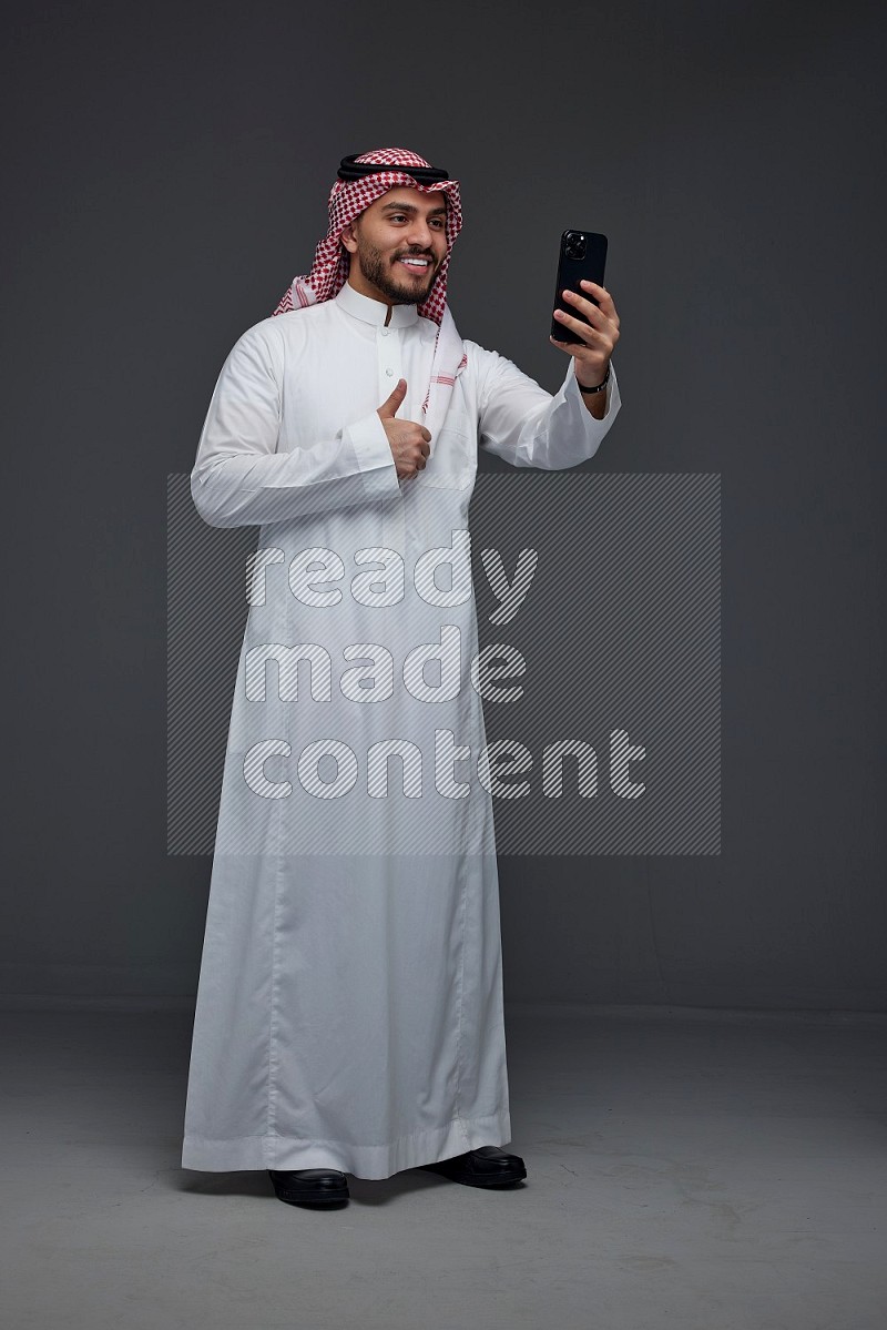 A Saudi man wearing Thobe and Shamgh taking selfie with his phone eye level on a gray background