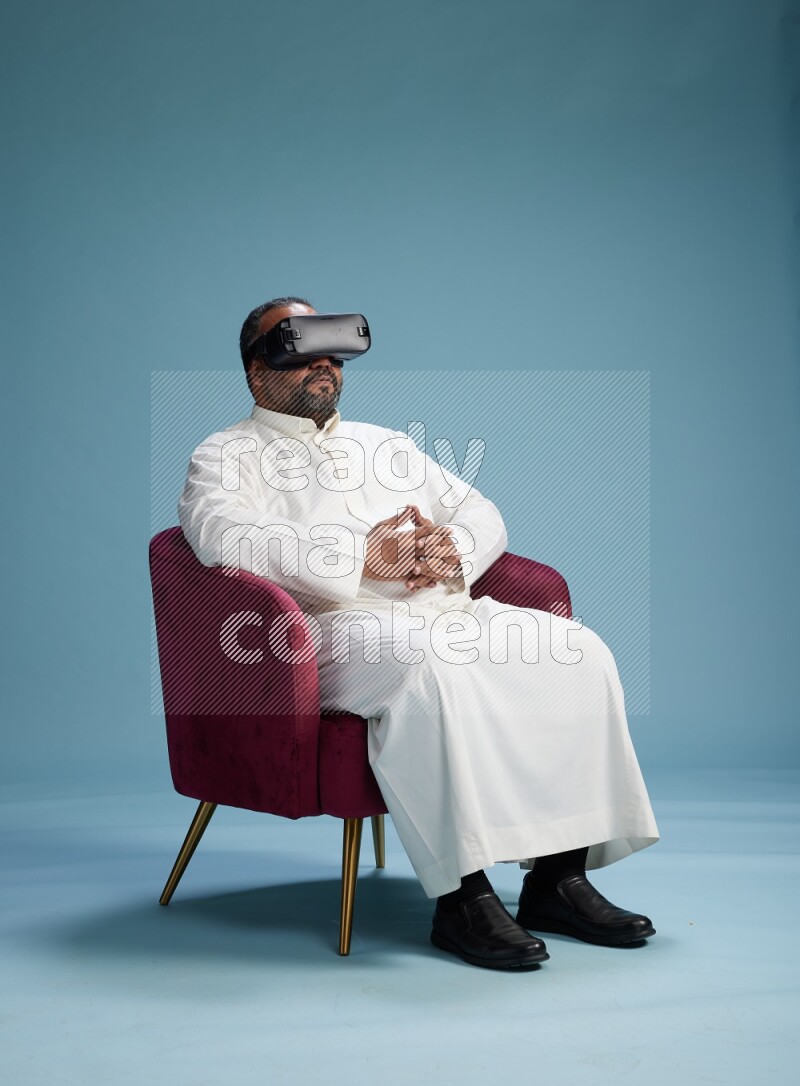 Saudi Man without shimag sitting on chair  gaming with VR on blue background