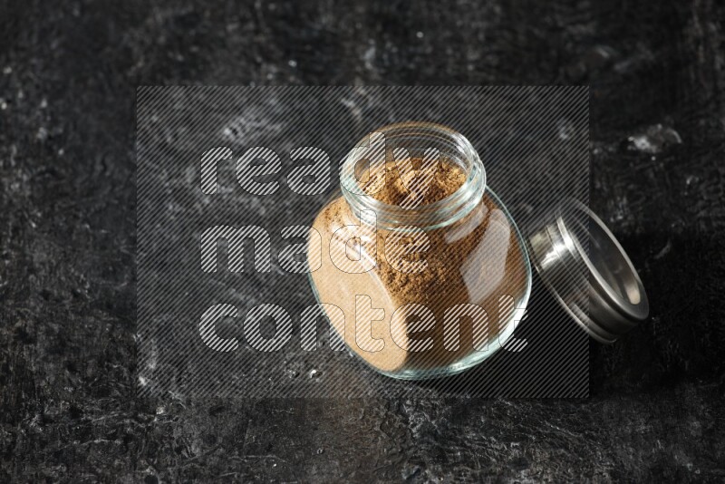 A glass spice jar full of allspice powder on a textured black flooring