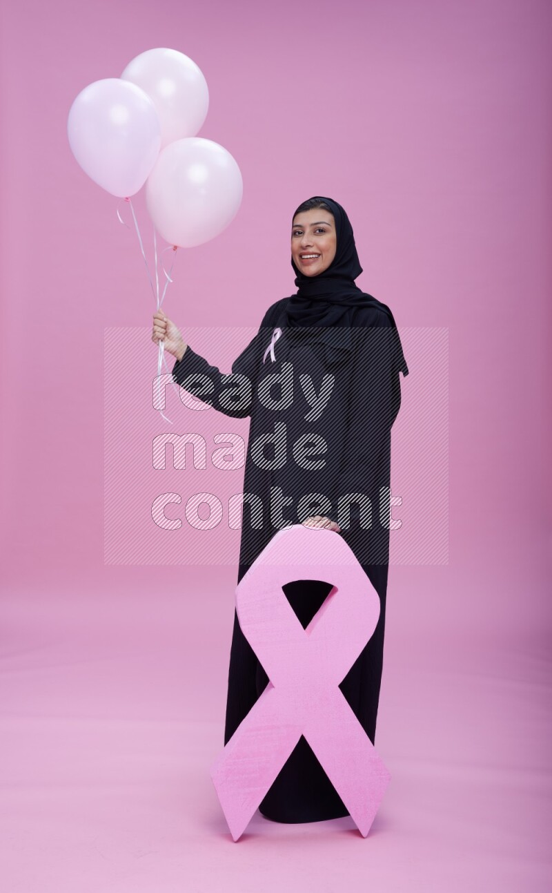 Saudi woman wearing pink ribbon on Abaya standing holding balloons and awareness sign on pink background