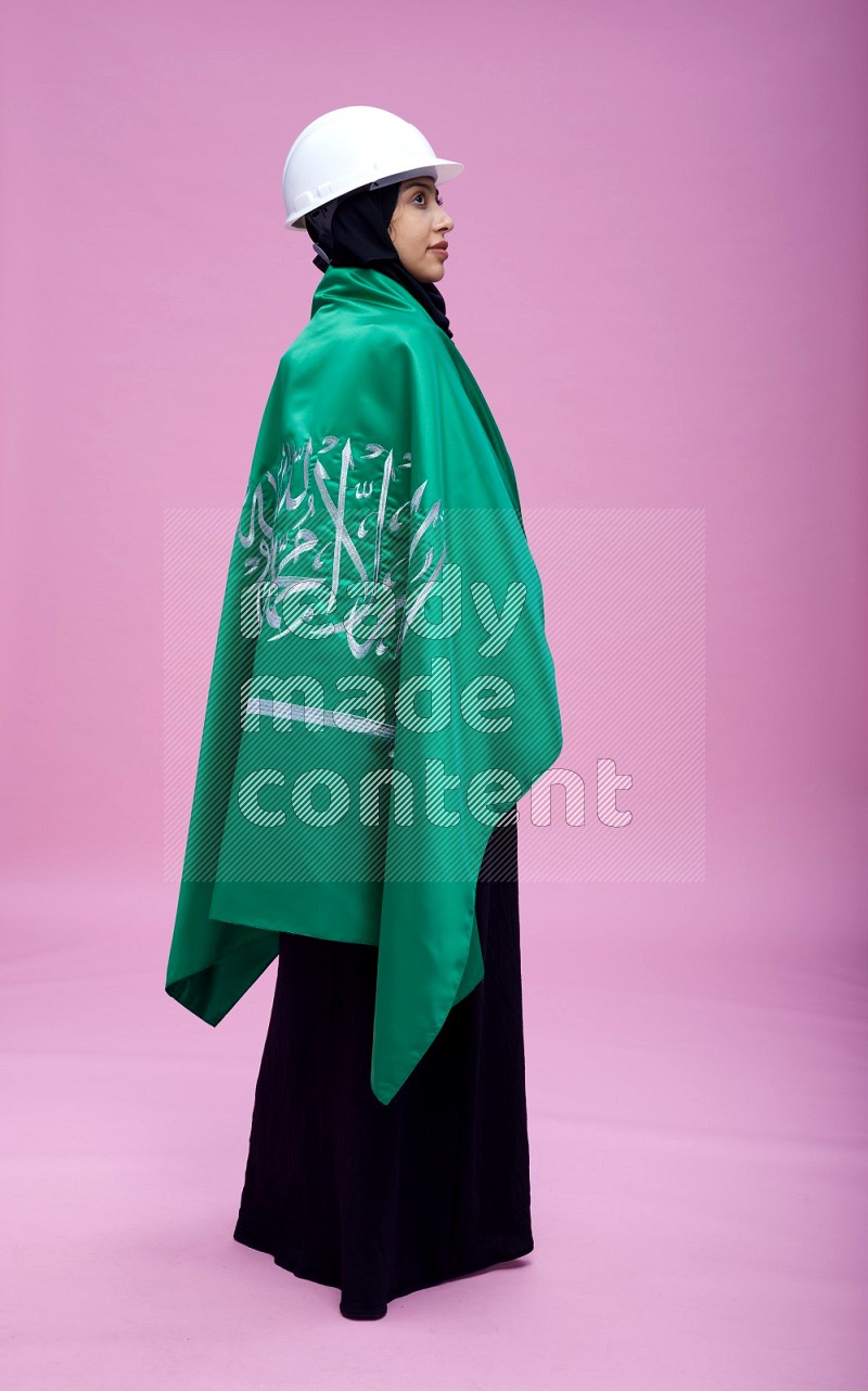 Saudi woman wearing Abaya with engineer vest and helmet standing holding Saudi flag on pink background