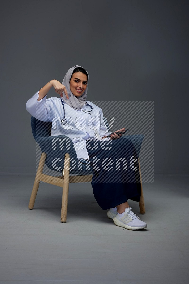 A doctor wearing a light gray head scarf setting on a dark grey chair and using a phone eye level on a grey background