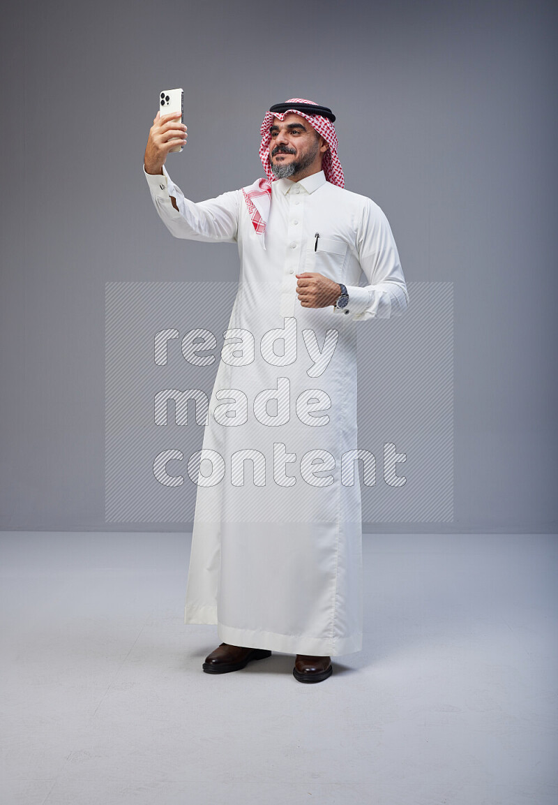 Saudi man Wearing Thob and red Shomag standing taking selfie on Gray background