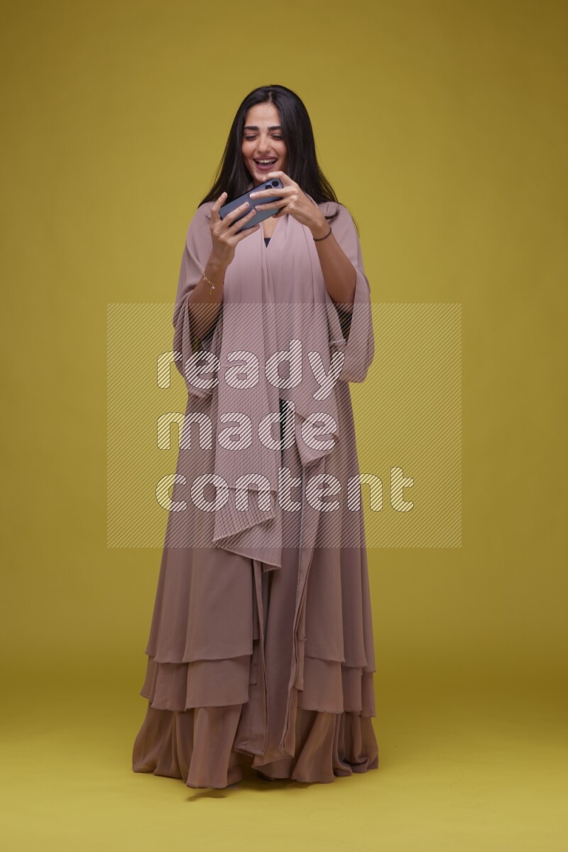 A woman Playing a Game on her smartphone on a Yellow Background wearing Brown Abaya