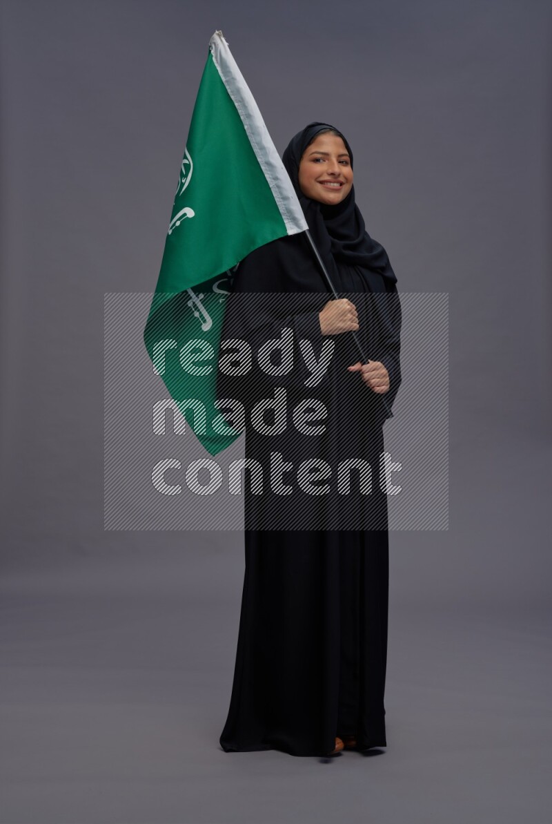 Saudi woman wearing Abaya standing holding Saudi flag on gray background