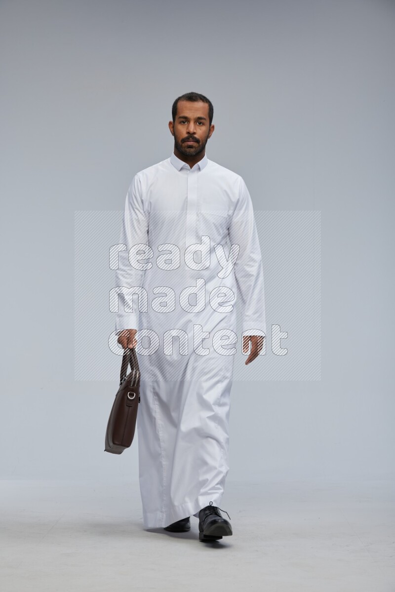 Saudi man wearing thob standing holding suitcase on gray background
