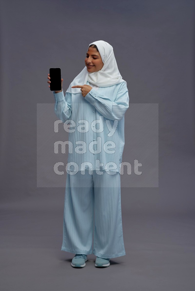 Saudi woman wearing hijab clothes standing showing phone to camera on gray background