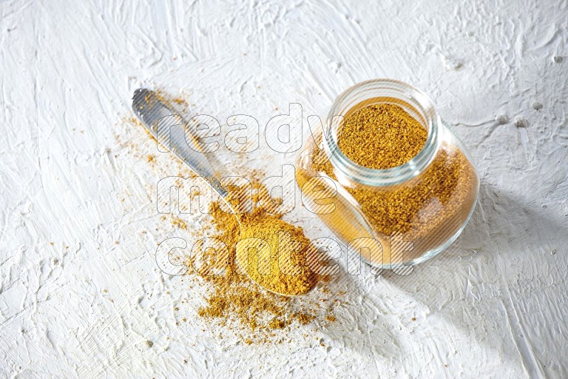 A glass spice jar and metal spoon full of turmeric powder on textured white flooring