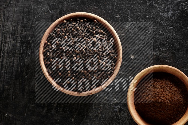 2 wooden bowls full of cloves powder and whole cloves on a textured black flooring