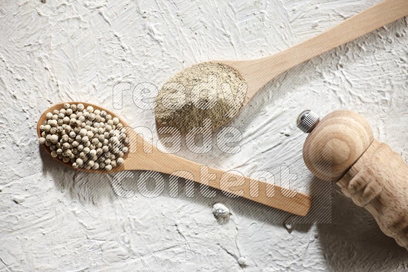 2 wooden spoons one full of white pepper powder and the other with pepper beads and a wooden pepper grinder on textured white flooring