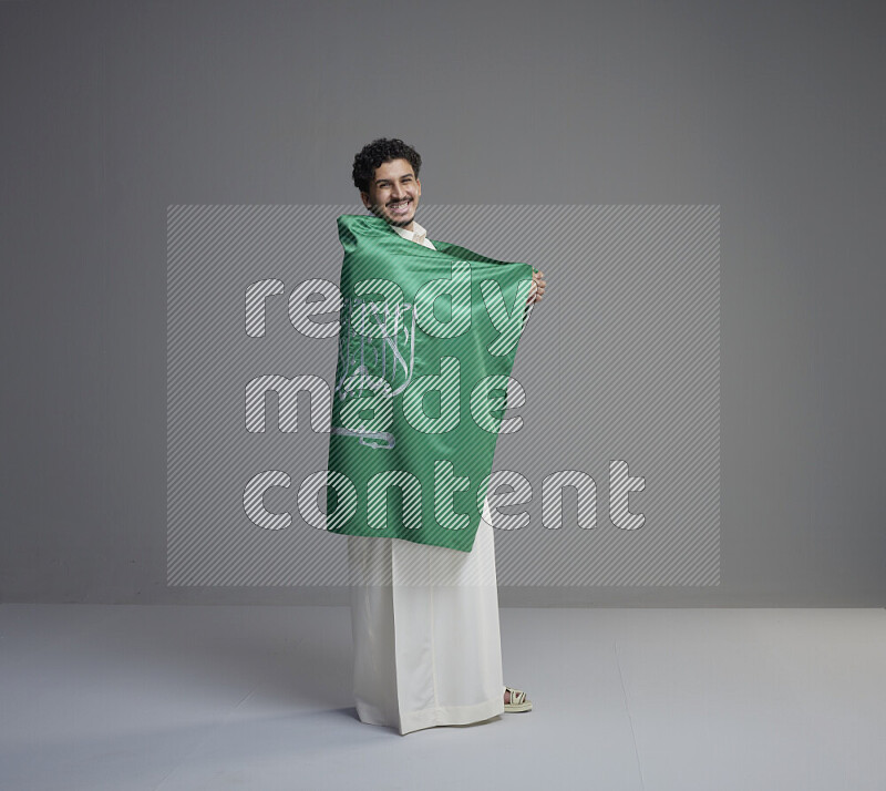 A Saudi man standing wearing thob wrapping big Saudi flag on gray background