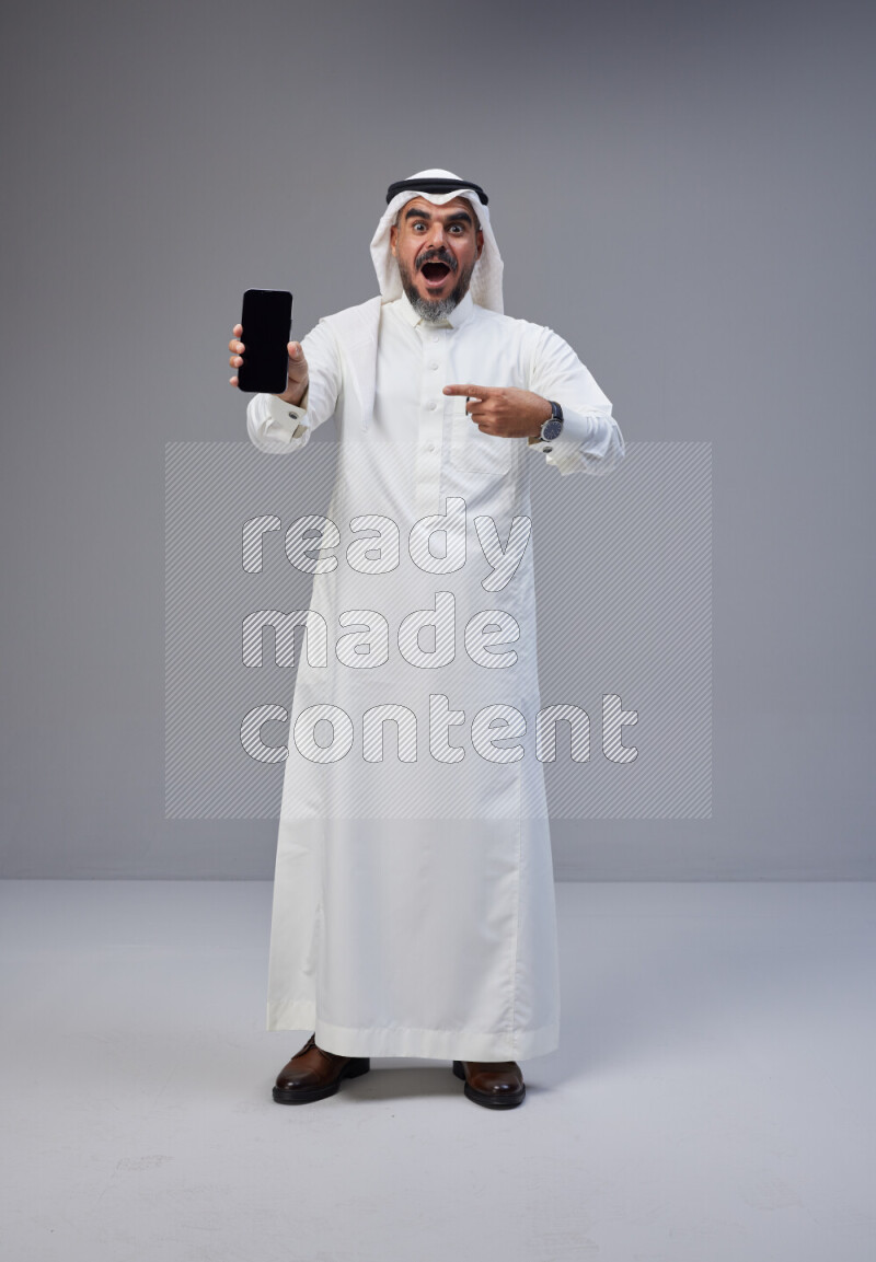 Saudi man Wearing Thob and white Shomag standing showing phone to camera on Gray background