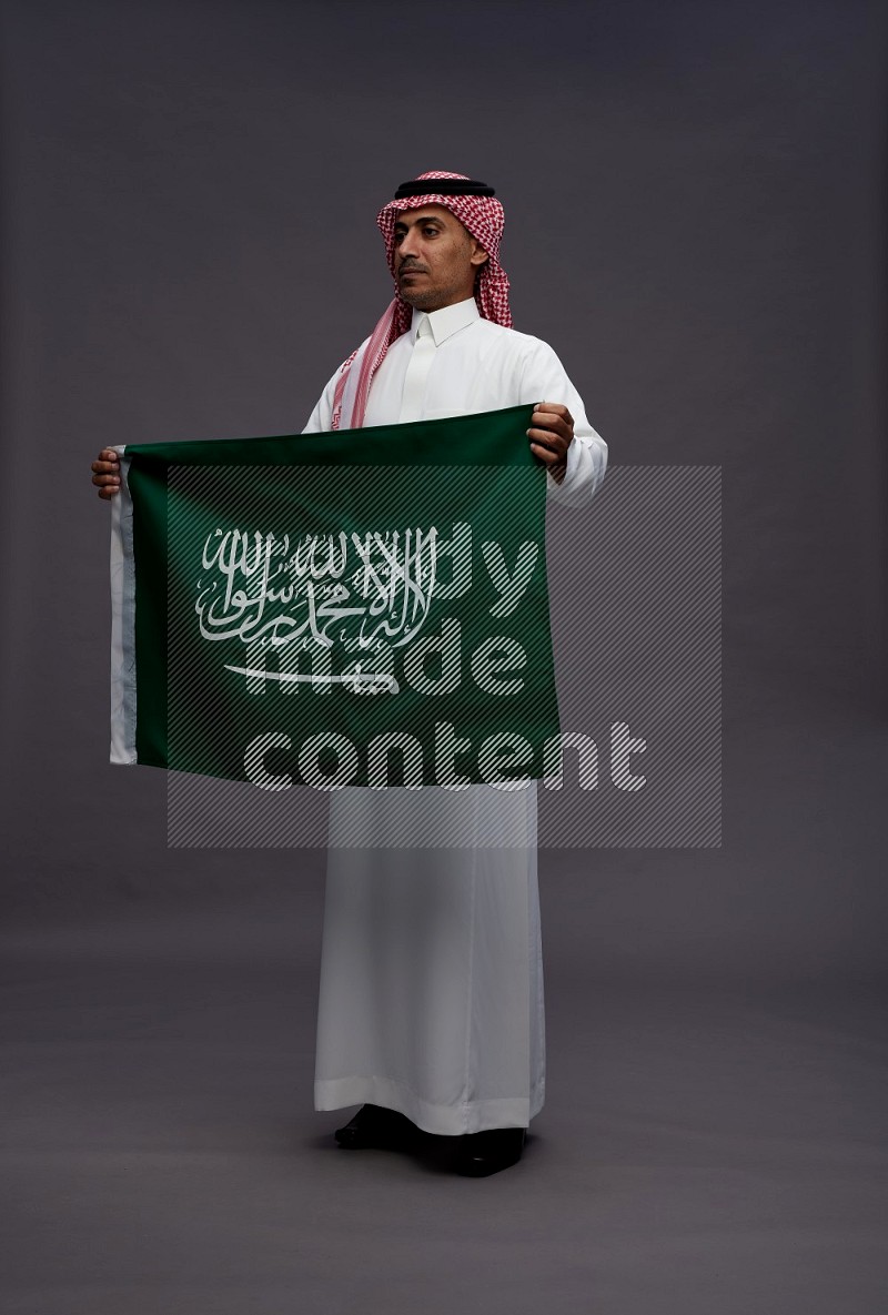 Saudi man wearing thob and shomag standing holding Saudi flag on gray background