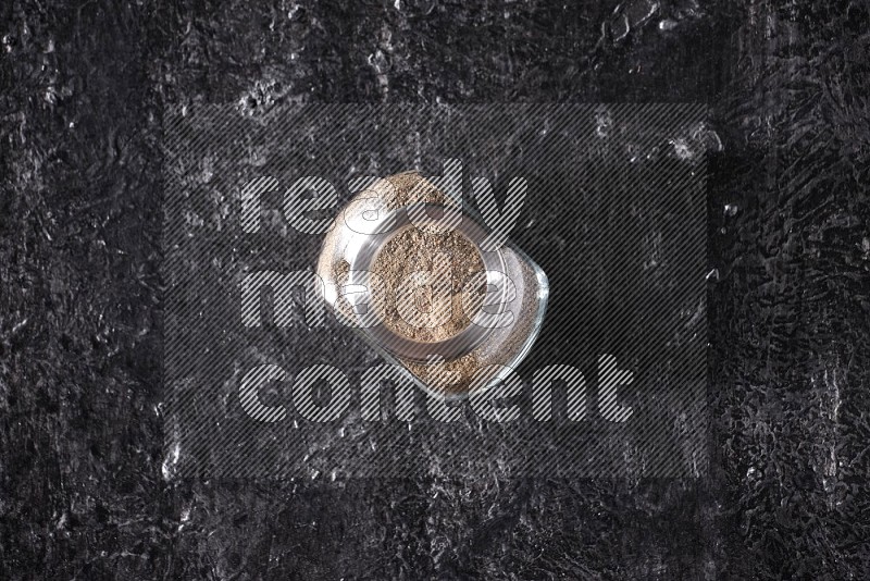 A glass spice jar full of black pepper powder on textured black flooring