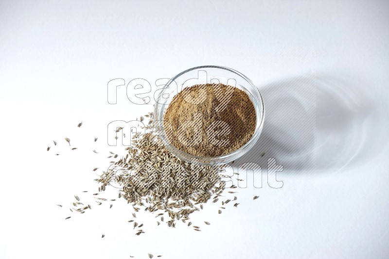 A glass bowl full of cumin powder with cumin seeds beneath it on white flooring