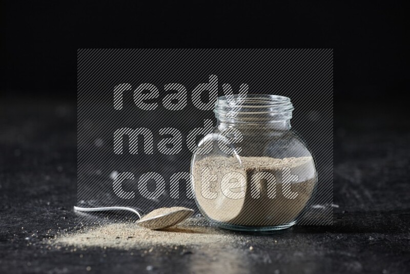 Herbal glass jar and metal spoon full of white pepper powder on textured black flooring