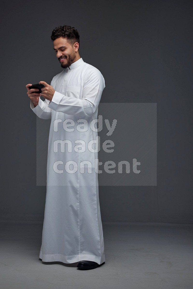 A Saudi man wearing Thobe and playing using his phone horizontally playing video games while standing and making different poses eye level on a gray background