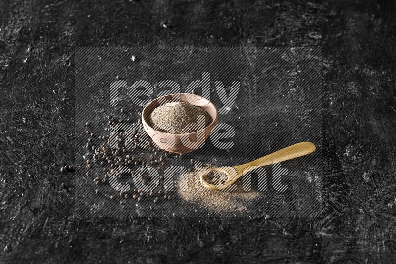 بودرة الفلفل الأسود في وعاء خشبي وملعقة خشبية مع الحبوب بجانبهم علي خلفية سوداء