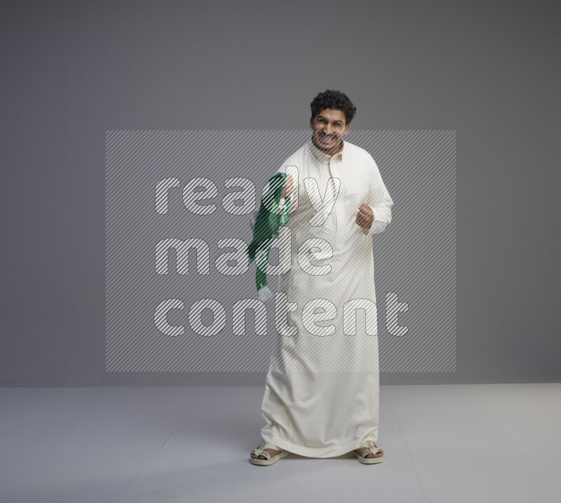 A Saudi man standing wearing thob holding Saudi flag scarf on gray background