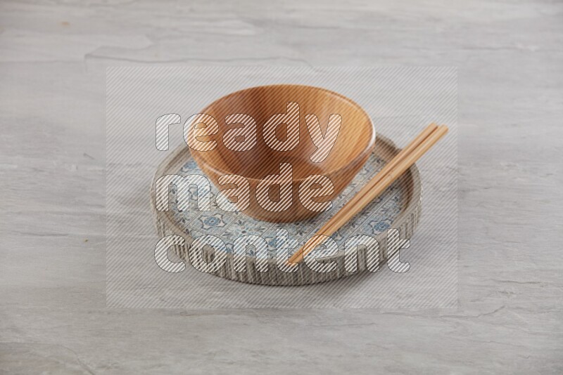 brown wood round bowl on top of multi color round ceramic plate and chopsticks, on grey textured countertop