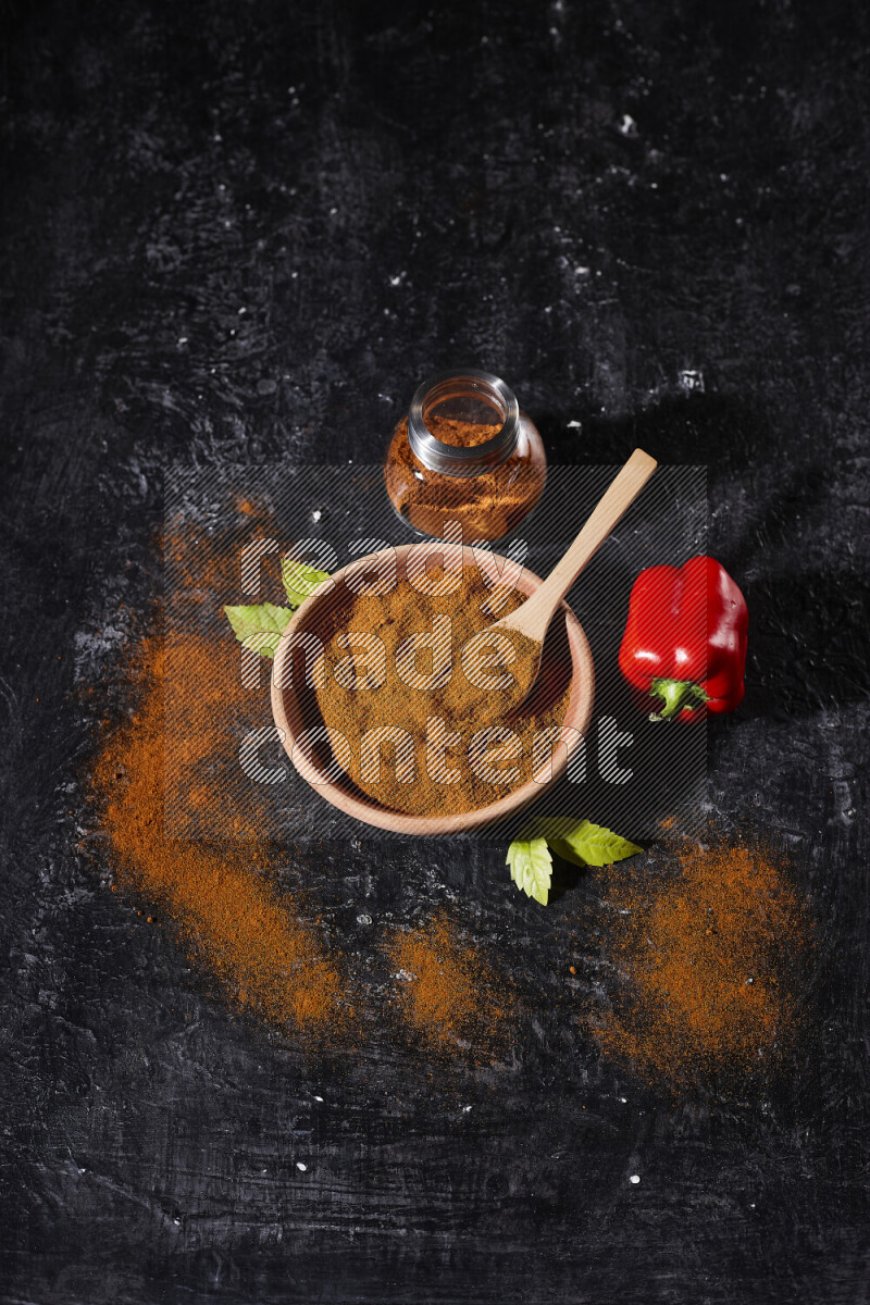 A wooden bowl full of ground paprika powder with a glass jar beside it and a red bell pepper on black background