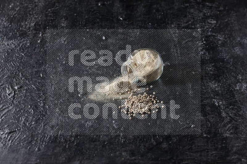 A flipped herbal glass jar full of white pepper powder with spilled powder and pepper beads on textured black flooring