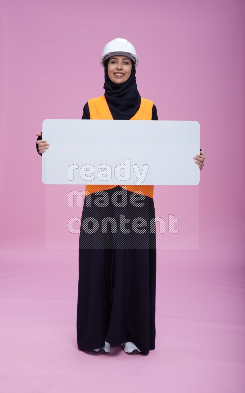Saudi woman wearing Abaya with engineer vest and helmet standing holding board on pink background